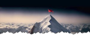 Mountain peak covered in white snow, with a trail to the pinnacle where a red flag sits.